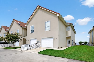 This photo shows a two-story, beige townhouse with a red-tiled roof, featuring a double garage and a small fenced front area with a lawn. The property is part of a neat, well-maintained community, near water, as suggested by the view in the background.
