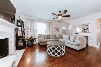 Living area featuring dark wood-style floors, ceiling fan, a stone fireplace, crown molding, and baseboards