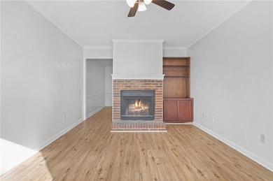 Unfurnished living room featuring light wood-style flooring, a ceiling fan, a brick fireplace, and crown molding