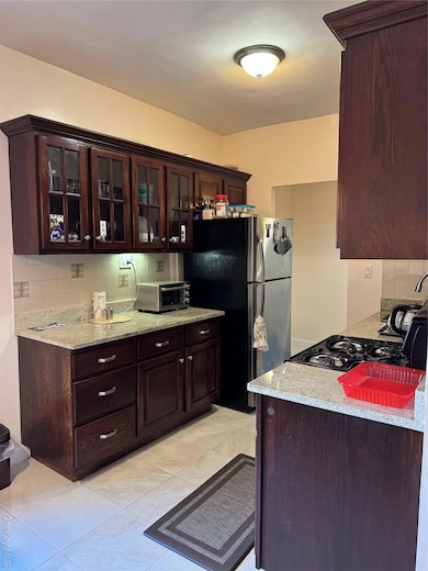 Kitchen featuring backsplash, glass insert cabinets, dark brown cabinetry, freestanding refrigerator, and light stone counters