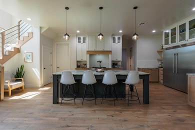 Kitchen featuring glass insert cabinets, stainless steel built in fridge, pendant lighting, an island with sink, and light stone counters