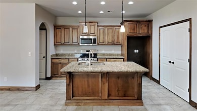 Kitchen featuring light stone counters, recessed lighting, appliances with stainless steel finishes, brown cabinetry, and an island with sink