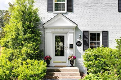 The front porch is blooming with color. The front entrance has a storm door, fluted columns, dentil molding, and handsome porch light.