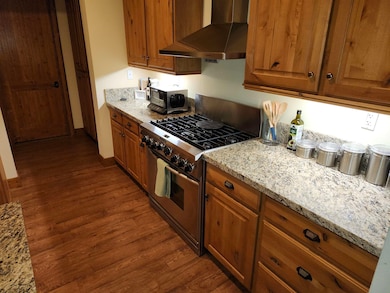 Kitchen featuring light stone countertops, wall chimney exhaust hood, gas stove, brown cabinetry, and dark wood-type flooring