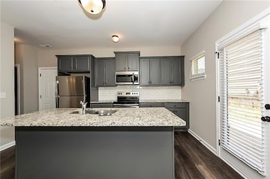 Kitchen featuring appliances with stainless steel finishes, backsplash, a kitchen island with sink, dark wood-type flooring, and light stone counters