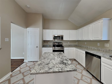 Kitchen with stainless steel appliances, inlaid floor details, light stone countertops, white cabinets, and lofted ceiling