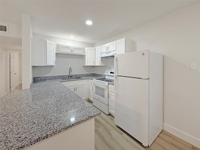 Modern kitchen with white cabinets and granite countertops. Features include a refrigerator, oven, and ample storage space. Bright and inviting with light wood flooring.