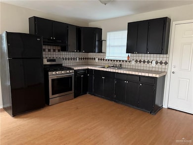 Kitchen featuring freestanding refrigerator, stainless steel range with gas stovetop, dark cabinets, ventilation hood, and tile countertops