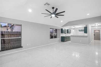 This spacious, open-concept living area features a modern ceiling fan, large windows for natural light, and connects seamlessly to a kitchen with stylish cabinetry. The neutral color palette and tile flooring create a clean, contemporary feel.