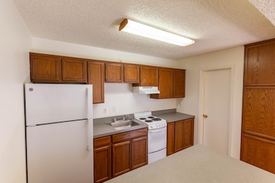 Kitchen featuring white appliances, brown cabinetry, a textured ceiling, under cabinet range hood, and light countertops