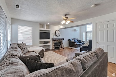 Living area featuring wood finished floors, built in shelves, a textured ceiling, a stone fireplace, and a ceiling fan