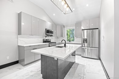 Kitchen with modern cabinets, stainless steel appliances, light stone counters, a kitchen island with sink, and lofted ceiling