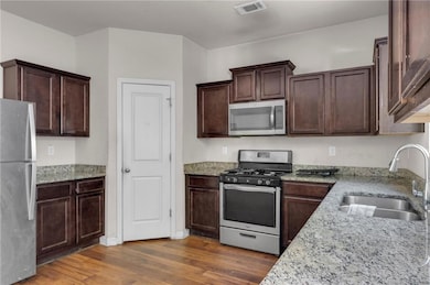 Kitchen with sink, dark wood-type flooring, light stone counters, dark brown cabinets, and appliances with stainless steel finishes
