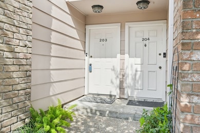 It features brick and siding walls, outdoor lighting, and a small concrete step with greenery nearby, creating a welcoming entryway.