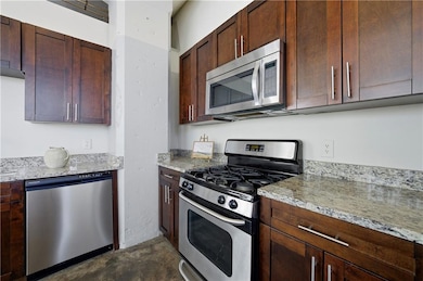 Kitchen with appliances with stainless steel finishes, light stone countertops, concrete floors, and dark brown cabinetry