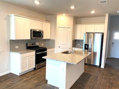 Kitchen with appliances with stainless steel finishes, white cabinets, light stone countertops, dark wood-type flooring, and a kitchen island with sink