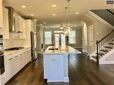 Kitchen with ceiling fan, light countertops, wall chimney range hood, white cabinetry, and an island with sink