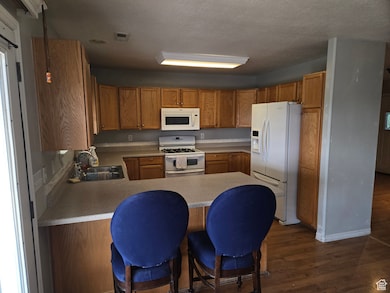 Kitchen featuring dark wood-style floors, white appliances, a peninsula, a breakfast bar, and brown cabinets
