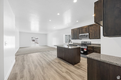 Kitchen featuring dark brown cabinets, a kitchen island with sink, open floor plan, stainless steel appliances, and light stone counters
