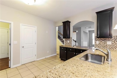 Kitchen featuring light tile patterned floors, backsplash, light stone countertops, arched walkways, and dark cabinets