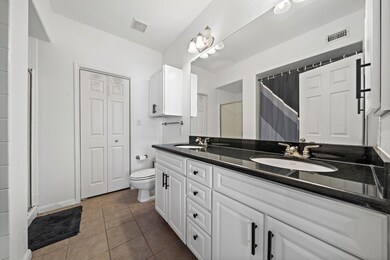 This bathroom features a modern design with a double vanity, black countertops, and white cabinetry. It includes a shower with a curtain, tiled flooring, and ample lighting, offering a clean and functional space.