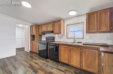 Kitchen with black appliances, under cabinet range hood, dark wood finished floors, brown cabinetry, and a decorative wall