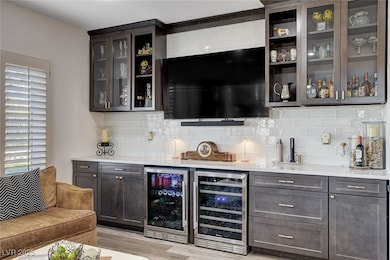 Indoor dry bar featuring wine cooler, light stone countertops, glass insert cabinets, and tasteful backsplash