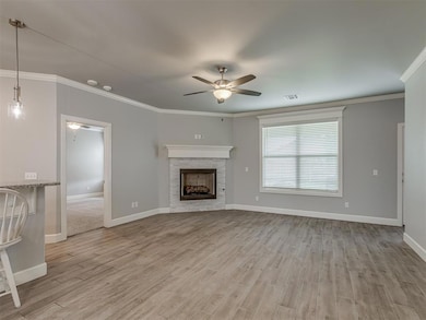 Unfurnished living room featuring a ceiling fan, light wood-type flooring, crown molding, baseboards, and a stone fireplace