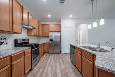 This modern kitchen features granite countertops, stainless steel appliances, and ample cabinet space. The open layout includes a large island with dual sinks and pendant lighting, complemented by wood-style flooring creating a warm and functional cooking space.