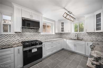 Main kitchen with backsplash, stainless steel appliances, light stone countertops, glass insert cabinets, and white cabinets