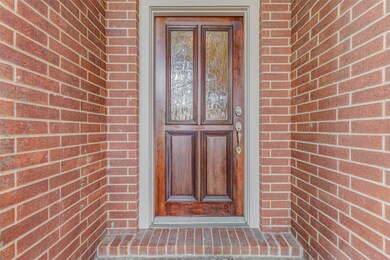 The welcoming wood front door is a true statement piece, boasting rich wood tones, decorative beveled glass panels, and an elegant brushed-nickel handle set. Nestled in a recessed brick entryway, it offers privacy and shelter, setting the stage for a refined interior. A transom window overhead adds natural light to the foyer inside while reinforcing the home’s classic design.