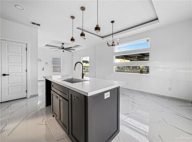 Kitchen featuring ceiling fan with notable chandelier, a tray ceiling, sink, pendant lighting, and an island with sink