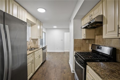 Kitchen with appliances with stainless steel finishes, under cabinet range hood, dark stone countertops, dark wood-style flooring, and backsplash