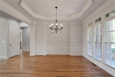Unfurnished dining area with a tray ceiling, a chandelier, light wood-type flooring, ornate columns, and a decorative wall