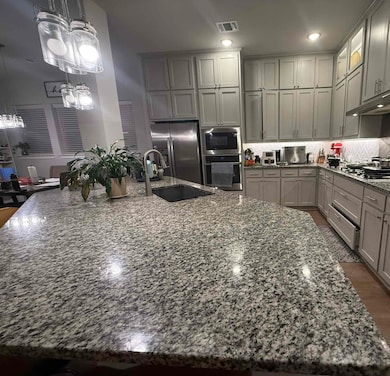 Kitchen featuring gray cabinets, light stone counters, hanging light fixtures, appliances with stainless steel finishes, and a center island with sink