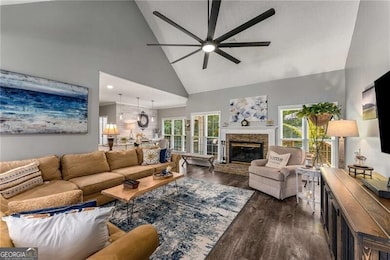 Living room featuring a ceiling fan, high vaulted ceiling, dark wood-style flooring, a stone fireplace, and healthy amount of natural light