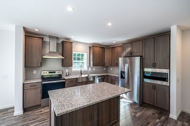Kitchen featuring stainless steel appliances, dark brown cabinetry, wall chimney exhaust hood, light stone counters, and dark wood-style flooring