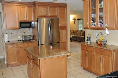Kitchen with freestanding refrigerator, backsplash, black microwave, light tile patterned floors, and light stone counters