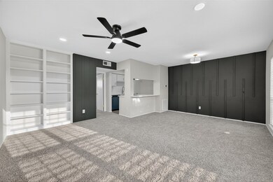 Spacious, modern living area with plush carpeting, built-in shelving, and a stylish black accent wall. Features recessed lighting, a ceiling fan, and an open layout connected to the kitchen.