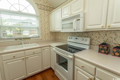 Kitchen with white appliances, a sink, wallpapered walls, dark wood-style flooring, and white cabinetry