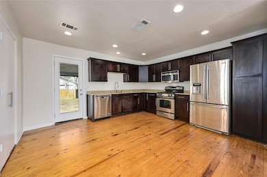 Kitchen with stainless steel appliances, dark brown cabinets, light wood-style flooring, recessed lighting, and light stone countertops