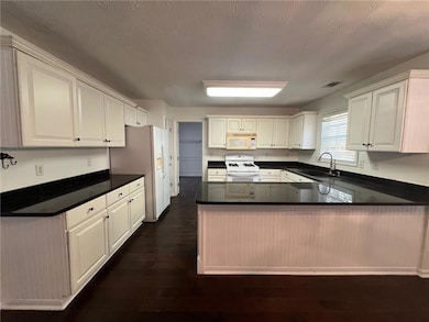 Kitchen with a peninsula, a textured ceiling, white appliances, dark wood-style floors, and dark stone countertops