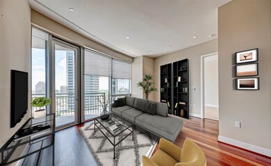 This modern living room features floor-to-ceiling windows with city views, sleek hardwood floors, and an open layout. It includes a contemporary gray sofa, a glass coffee table, and a tall bookshelf. Access to a balcony enhances the space with natural light.