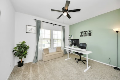 Designed for both productivity and comfort, this inviting home office features a soft neutral carpet, a calming green accent wall, and a large window with sheer curtains that fill the space with natural light. A modern desk and comfortable chair create an ideal workspace, while a cozy sofa offers a perfect spot to relax or read. Complete with a ceiling fan for year-round comfort, this space is as functional as it is stylish.