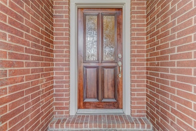 The welcoming wood front door is a true statement piece, boasting rich wood tones, decorative beveled glass panels, and an elegant brushed-nickel handle set. Nestled in a recessed brick entryway, it offers privacy and shelter, setting the stage for a refined interior. A transom window overhead adds natural light to the foyer inside while reinforcing the home’s classic design.