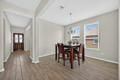 This photo features a bright dining area with wood-look tile flooring and neutral walls. It includes a chandelier above a four-seat dining table. Nearby, a hallway leads to a wooden front door with decorative glass panels, enhancing the home's inviting atmosphere.