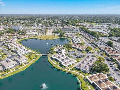 Expansive aerial view capturing the entire walking path around the palm-lined pond, featuring two fountains and lush, mature landscaping throughout the community. The palm tree-lined sidewalks and green spaces are clearly visible, along with the condo unit’s close proximity to the scenic pond and pathways. The view also highlights the convenient cul-de-sac parking area near the unit, showcasing the community’s accessible and well-planned layout.