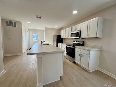 Kitchen with appliances with stainless steel finishes, white cabinets, light wood-type flooring, an island with sink, and light countertops