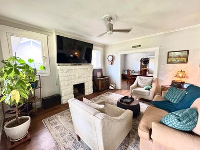 Living room featuring wood finished floors, a ceiling fan, ornamental molding, and a stone fireplace