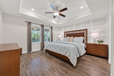 This spacious Primary bedroom features an elegant atmosphere, tray ceiling/crown molding, accent wall, recessed lighting and a ceiling fan. It includes large windows with curtains and blinds, a stylish bed with matching nightstands, and a dresser, all in warm wood tones. (Furniture available for purchase)